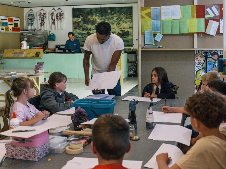 Children are sitting around a table, working together on an art activity. On the table are arts materials